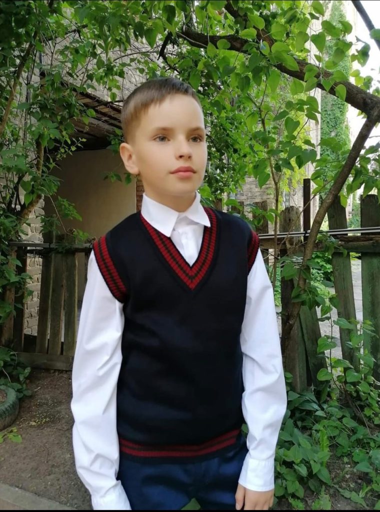 Zēns skolas formā: balts kreksl, tumši zila veste, tumši zilas bikses.